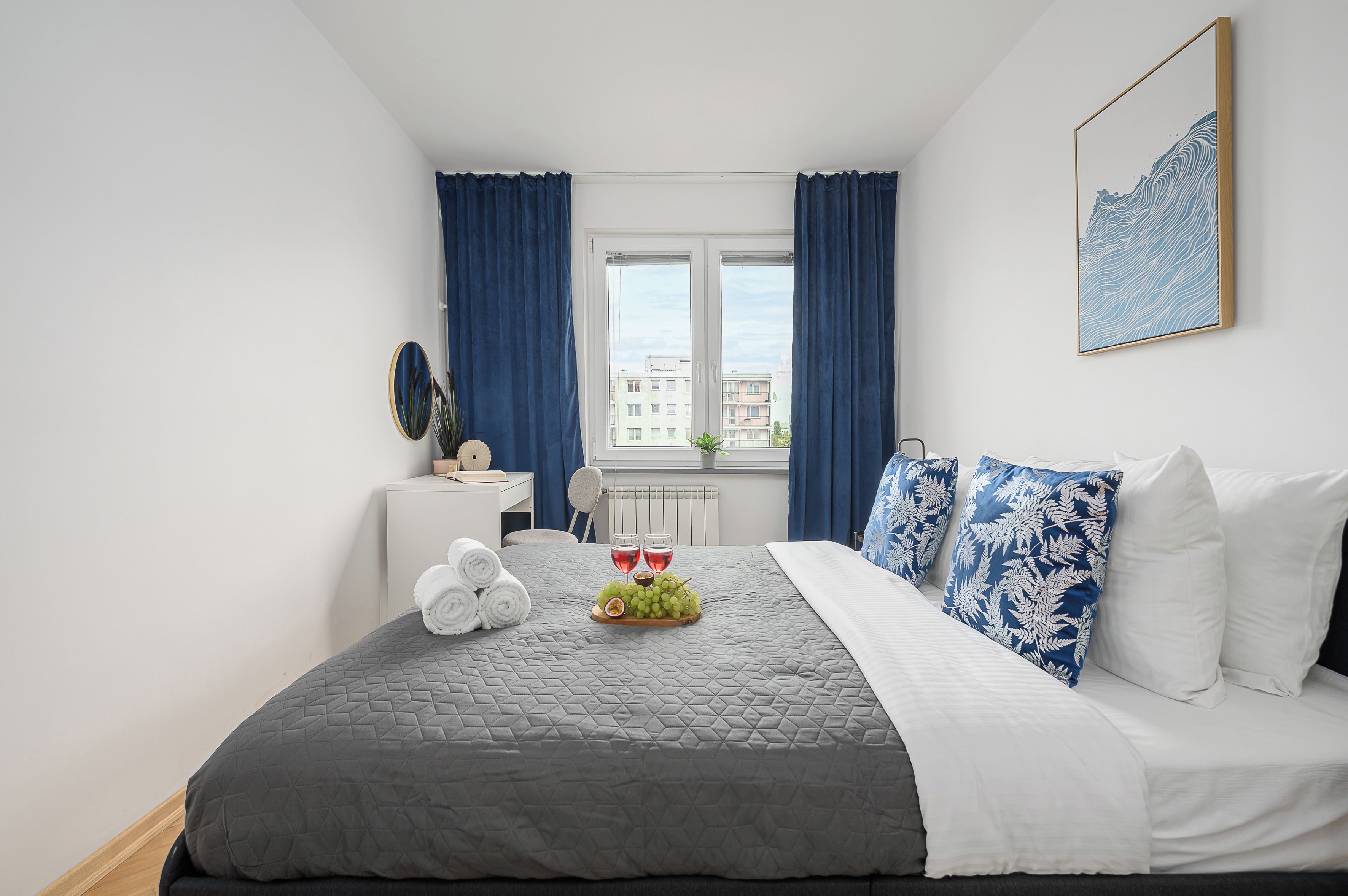 A hotel room with a bed, a desk, a chair, a window with curtains, and a wall-mounted picture.