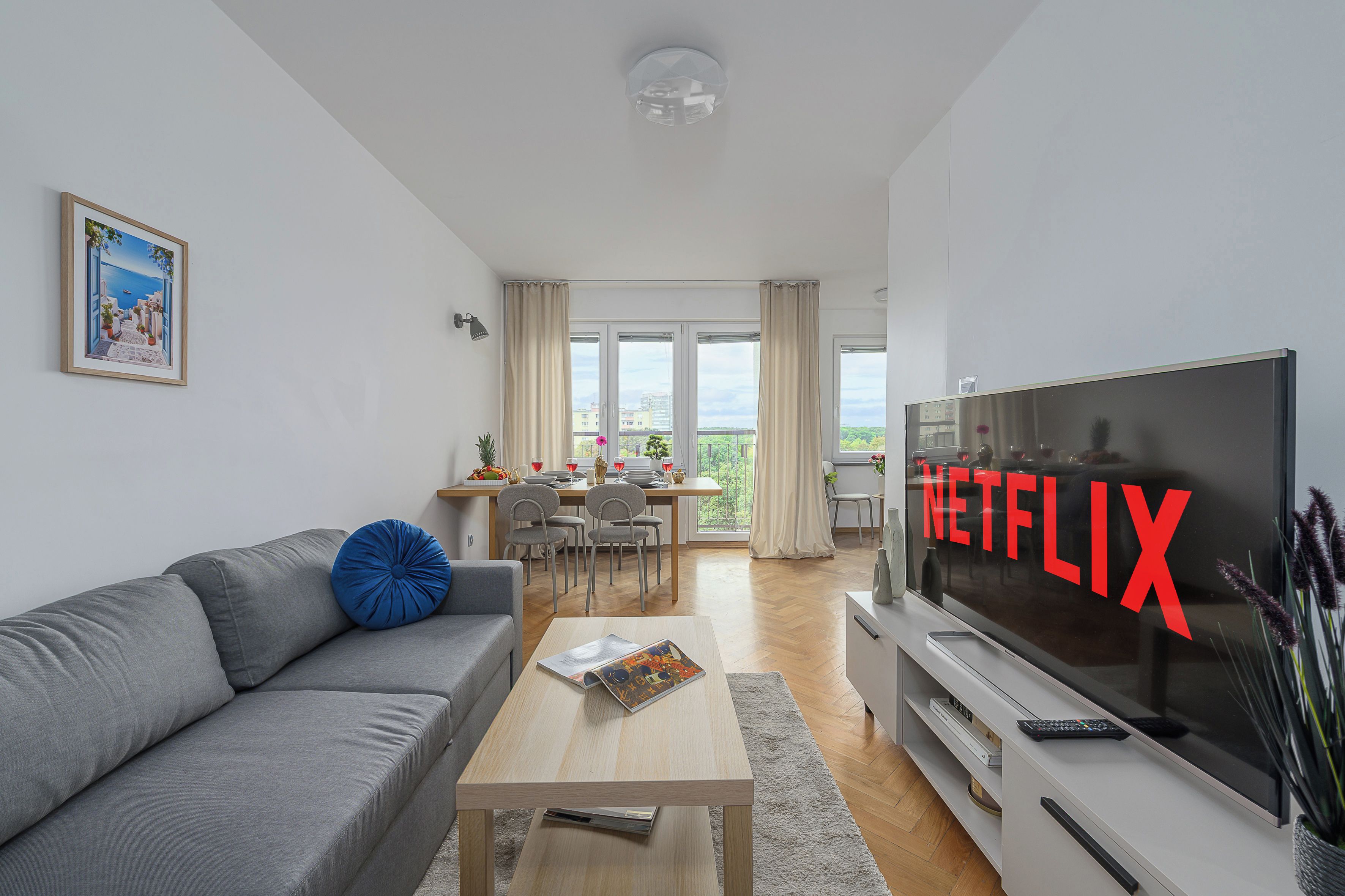 A modern living room with a grey sofa, a coffee table, a flat-screen TV, and a dining area with a table and chairs.