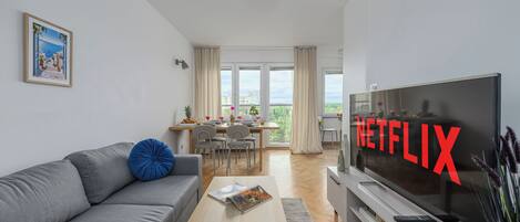 A modern living room with a grey sofa, a coffee table, a flat-screen TV, and a dining area with a table and chairs.