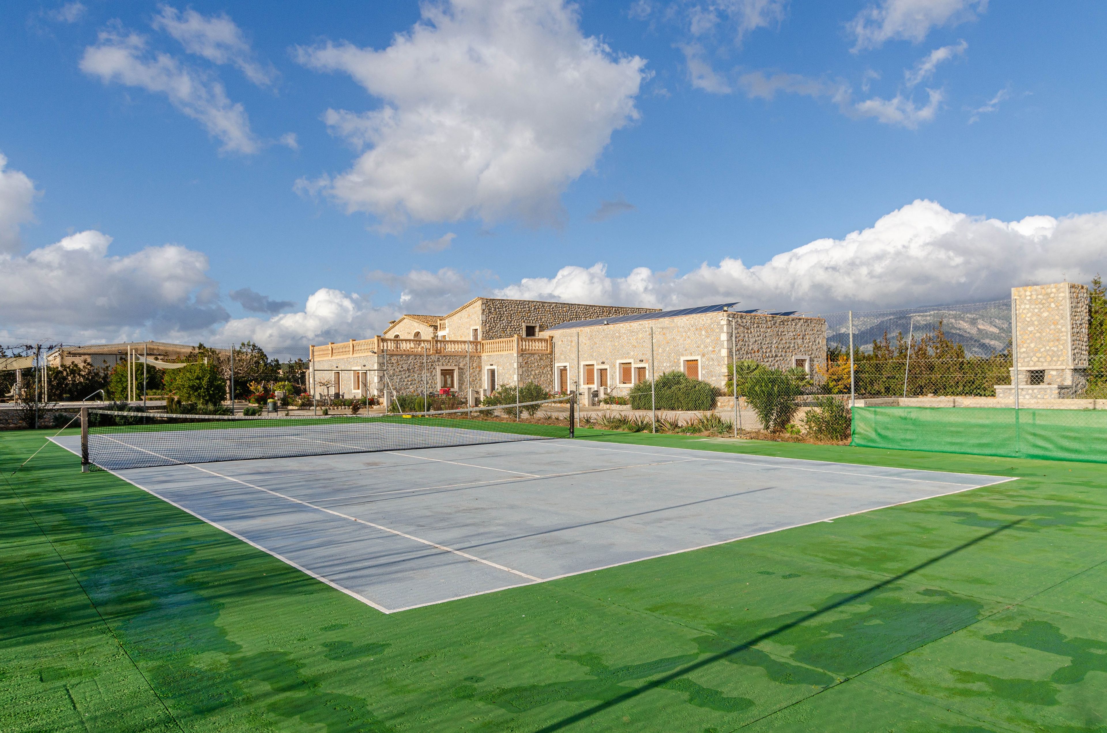 Cancha de tenis con red, rodeada por un área verde y un edificio de piedra con muro de piedra.