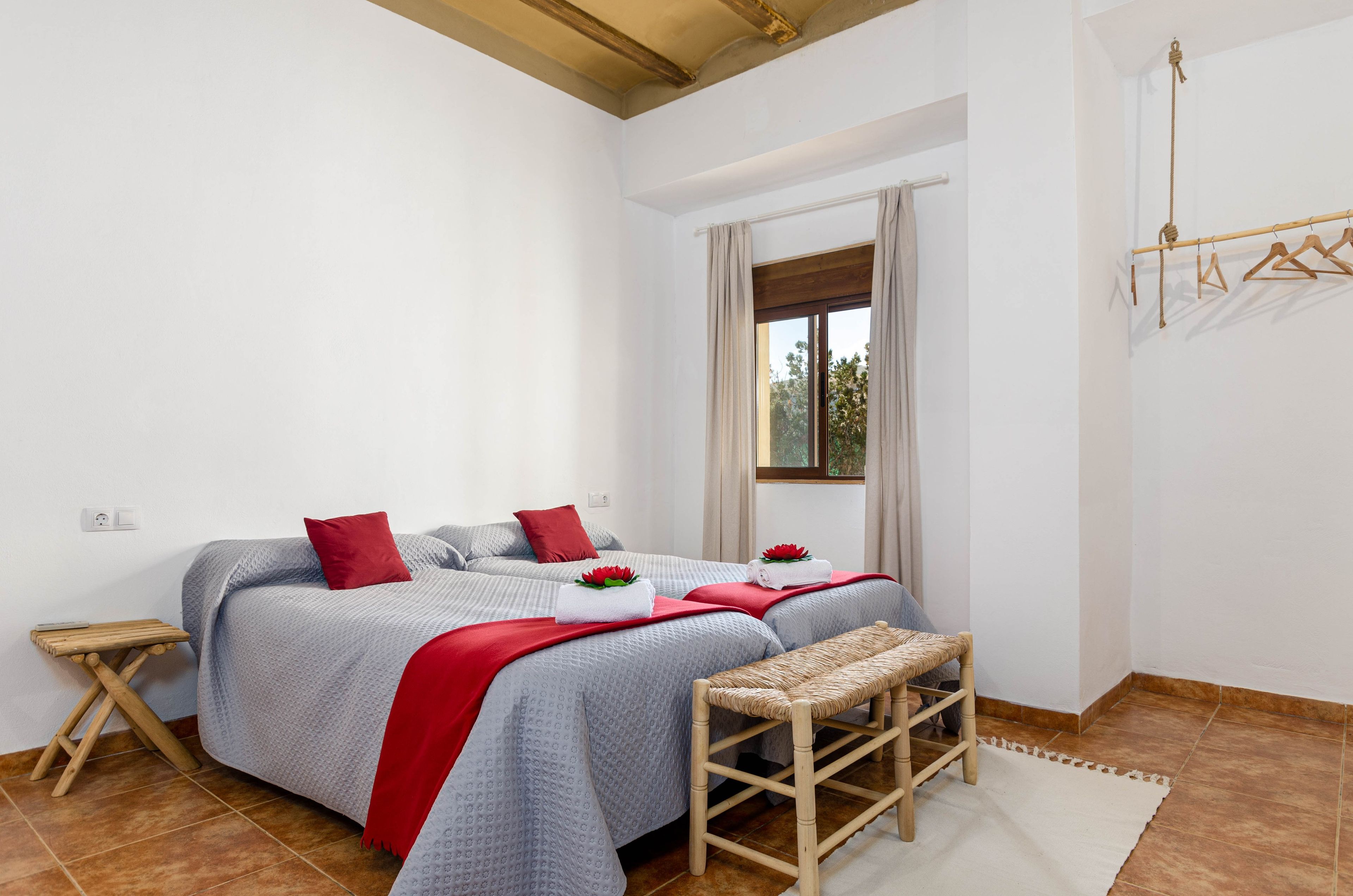 Una cama doble con una colcha roja, un taburete de madera pequeño y una ventana con cortinas.