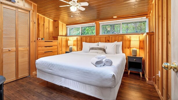 Een slaapkamer met houten lambrisering, een bed, twee bedlampen, een plafondventilator en een raam met jaloezieën.