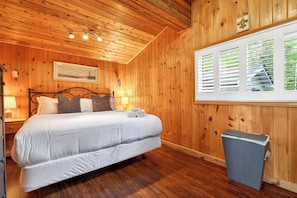 A wooden-paneled bedroom with a bed, bedside lamps, a framed picture, and a window with blinds. - Waterfront 3-bedroom cottage with hot tub and AC (Gravenhurst)