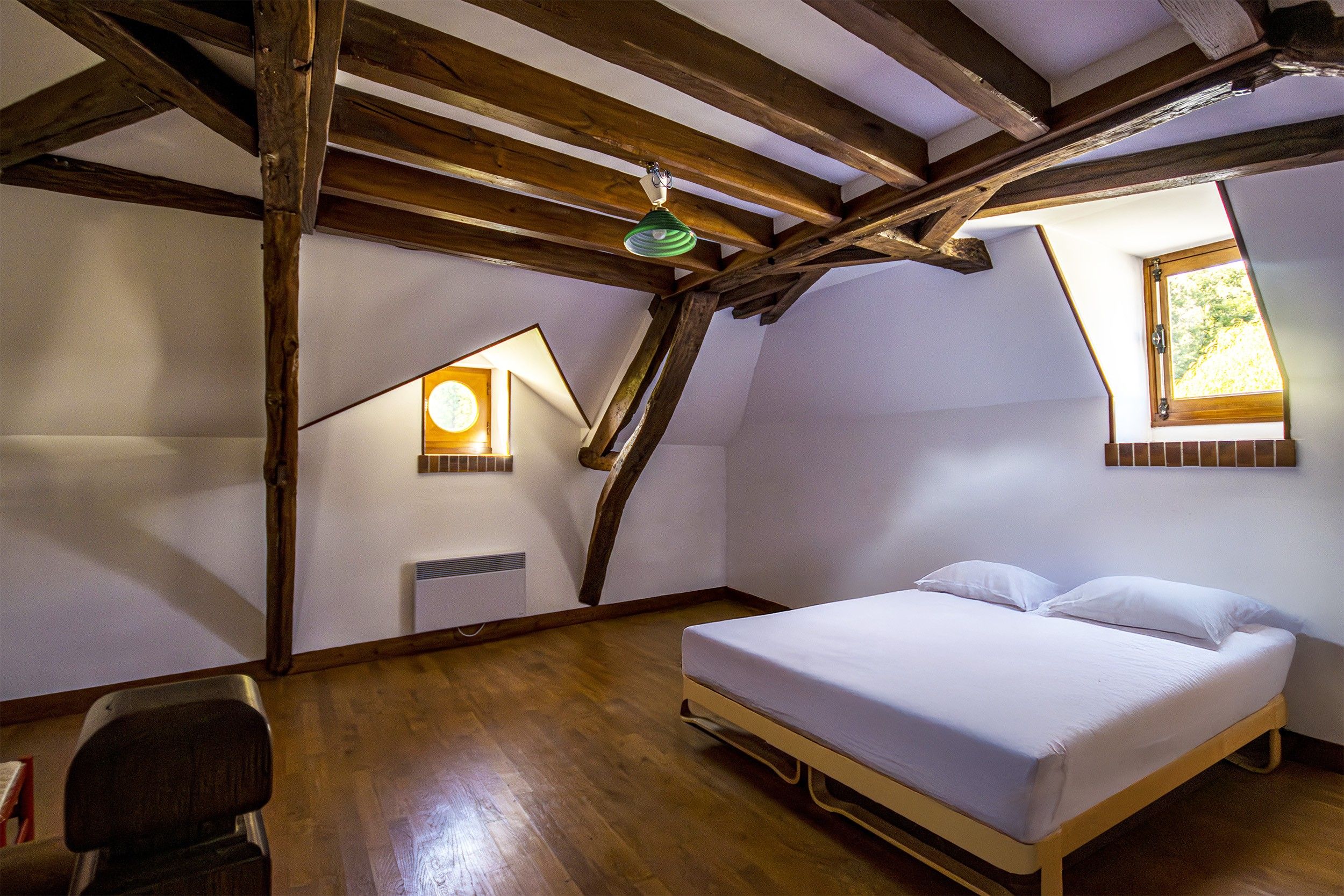 A bedroom with a wooden ceiling, a bed, a chair, a window, and a wall-mounted heater.