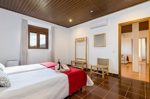 Een hotelkamer met twee bedden, een houten plafond, een airconditioner, een raam met gordijnen, een spiegel en een houten stoel.