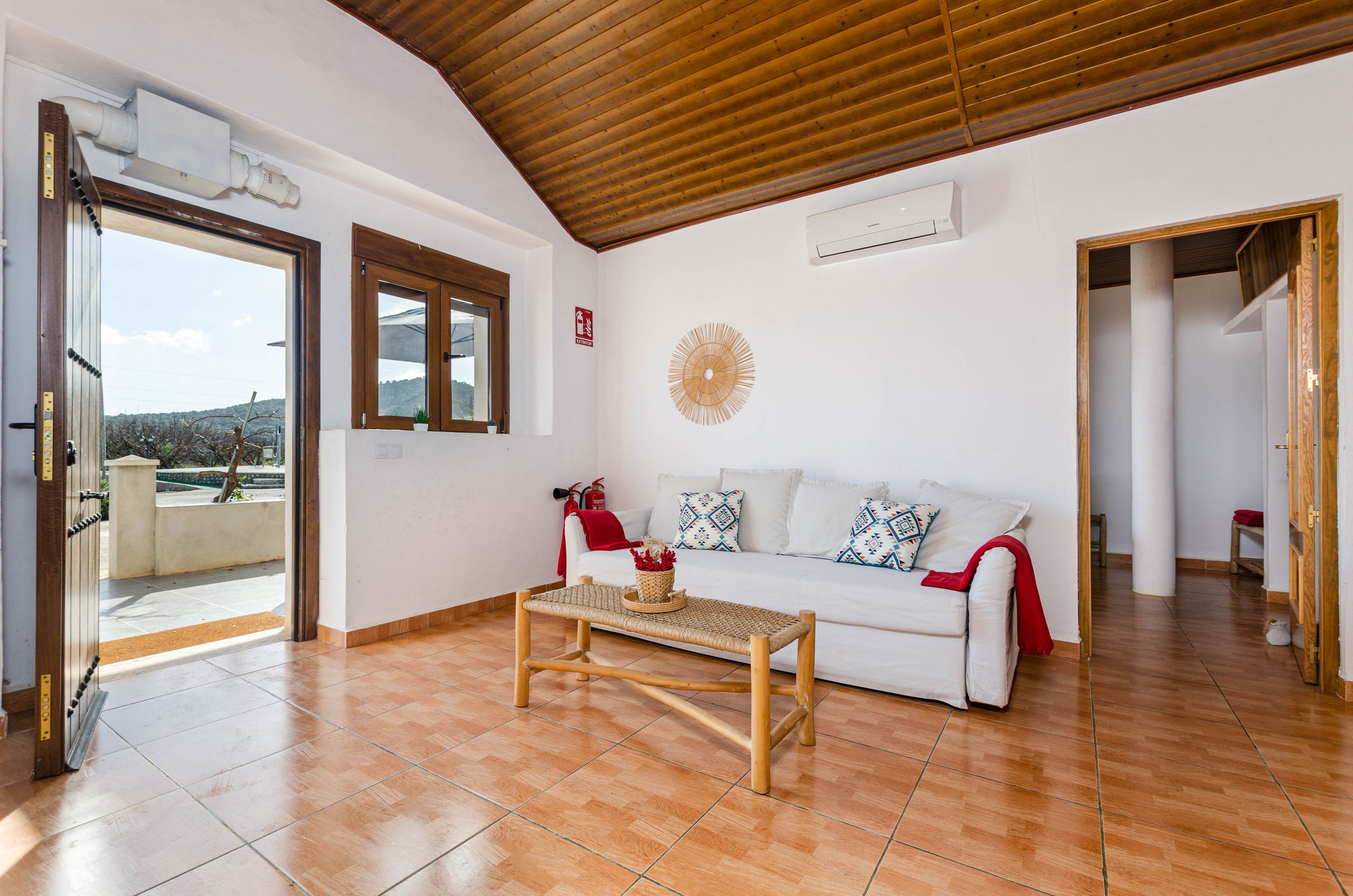 Amplia sala de estar con techo de madera, un sofá blanco con cojines estampados, una mesa de centro tejida y vistas a través de un ventanal abierto.