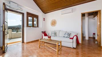 Amplia sala de estar con techo de madera, un sofá blanco con cojines estampados, una mesa de centro tejida y vistas a través de un ventanal abierto.