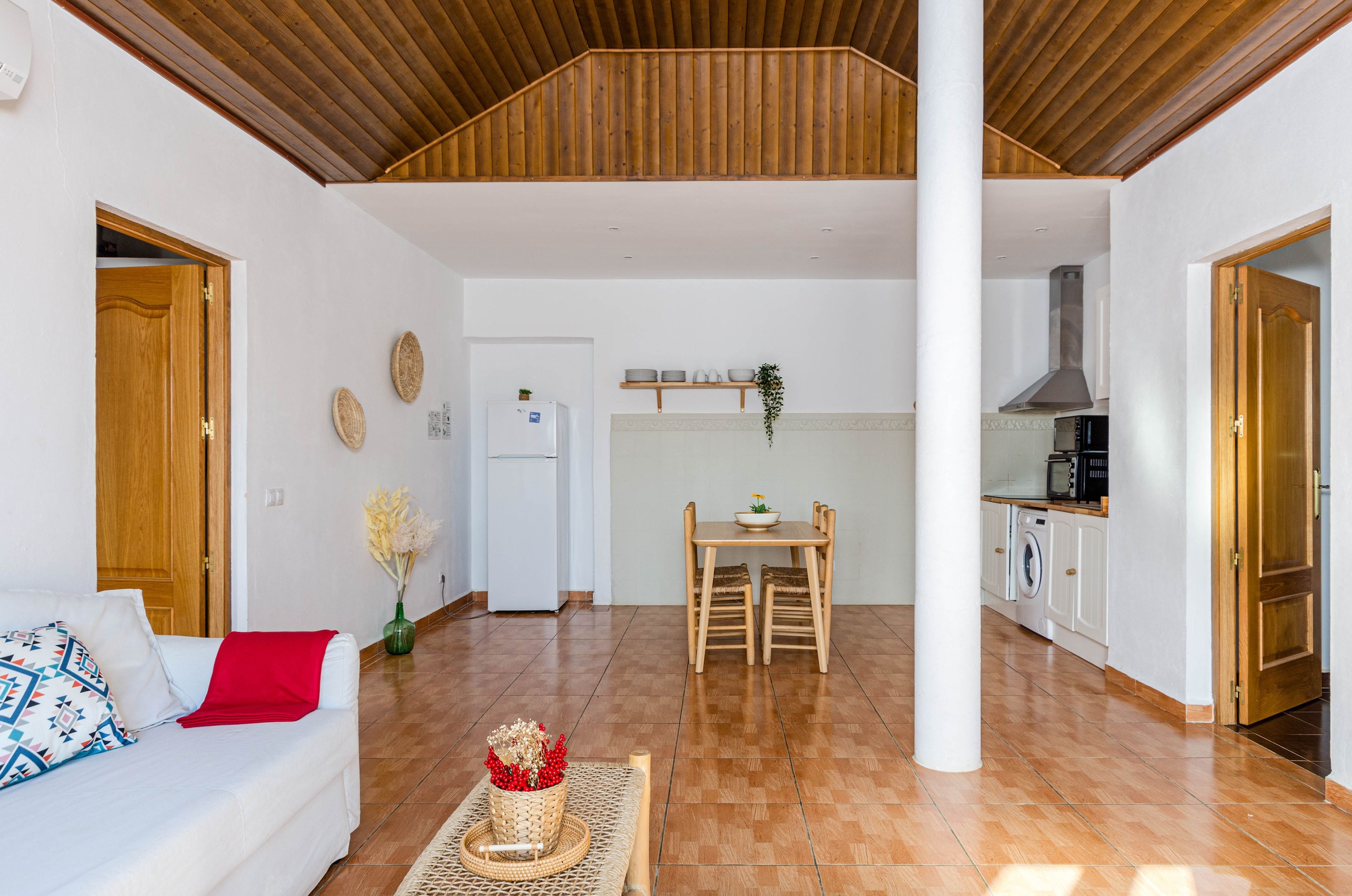 Amplia sala de estar con techo de madera, una mesa de comedor con sillas, una cocina con estufa y microondas, y un sofá blanco con un cojín rojo.