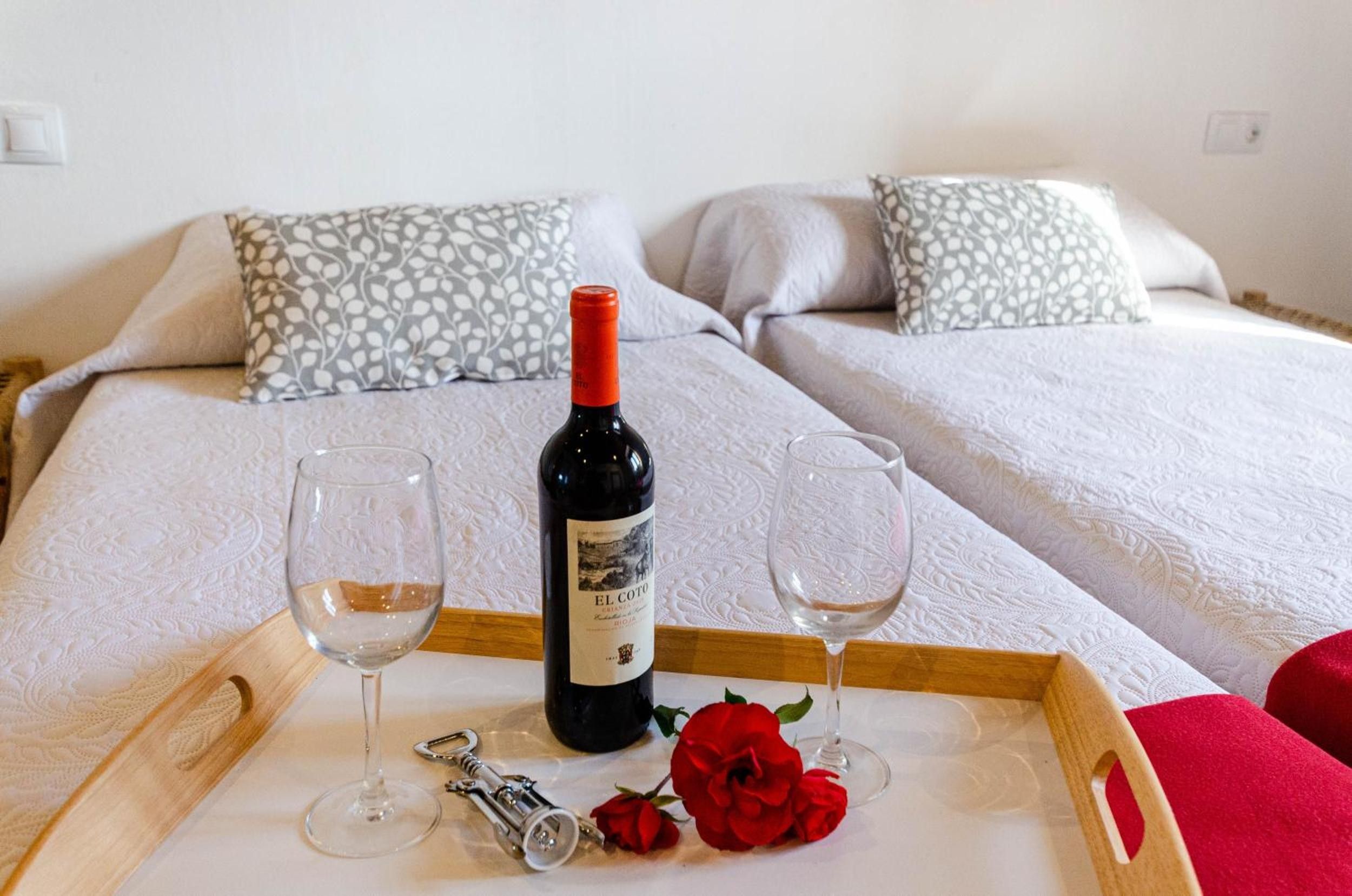 A bed with two pillows, a wine bottle, two glasses, a corkscrew, and a rose on a tray.