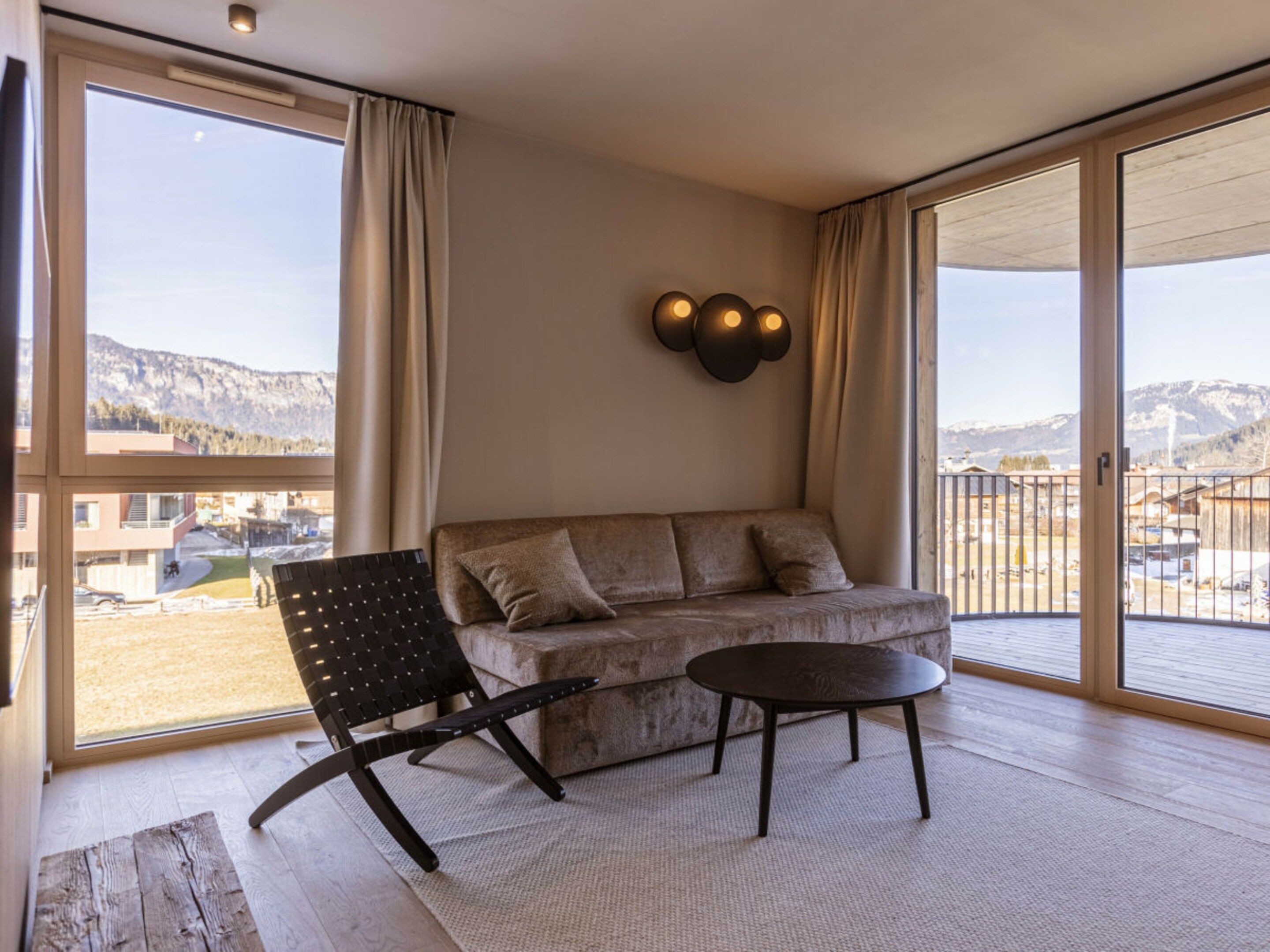 Een moderne woonkamer met een bank, een klein tafeltje en een stoel. Er is een groot raam met uitzicht op gebouwen en bergen.