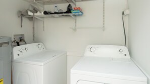 Une buanderie équipée de deux machines à laver et de deux sèche-linge à chargement frontal, tous deux de couleur blanche, d’une étagère contenant des produits de lessive et d’une prise électrique au mur.