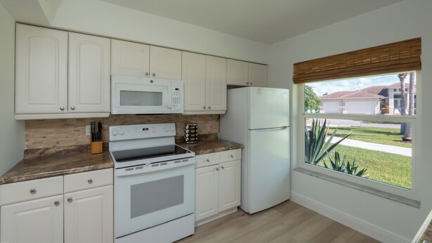 A kitchen with white cabinets, a microwave, a stove, a refrigerator, and a window with a view of a residential area. - Beautiful 3-bedroom, 2-bathroom waterfront home sits right on the canal! 1131 Fern Court (Marco Island)