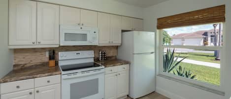 A kitchen with white cabinets, a microwave, a stove, a refrigerator, and a window with a view of a residential area.