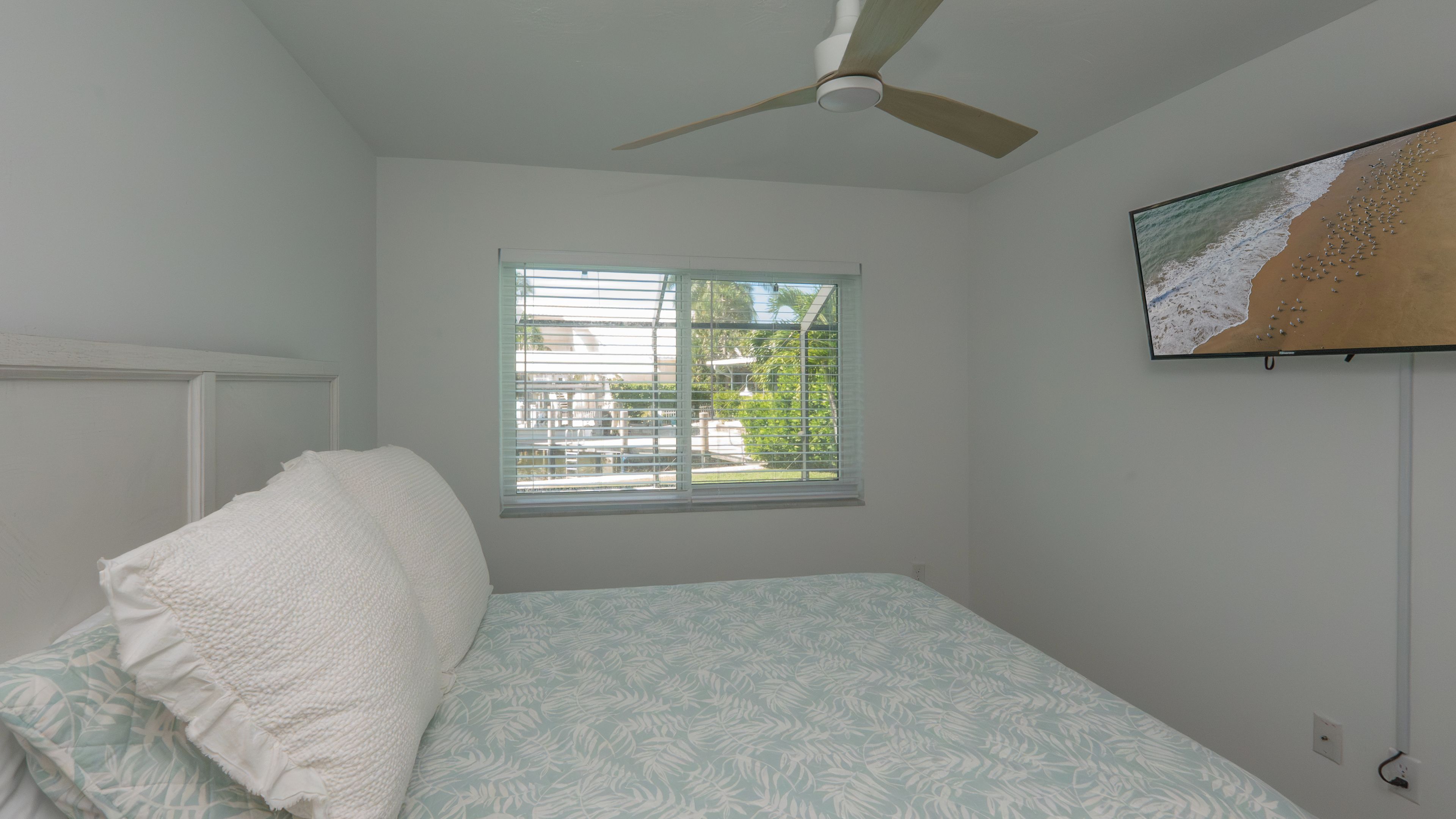A bedroom with a bed, a ceiling fan, a window with blinds, and a beach-themed wall hanging.