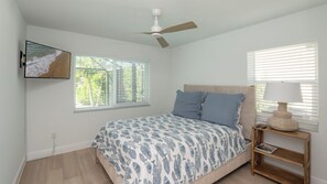 A bedroom with a bed, a ceiling fan, a nightstand with a lamp, a wall-mounted TV, and a window with blinds. - Beautiful 3-bedroom, 2-bathroom waterfront home sits right on the canal! 1131 Fern Court (Marco Island)