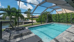 Une piscine dotée d’un espace couvert, de chaises longues et offrant une vue sur un quartier résidentiel avec des maisons et des palmiers.