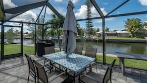 An outdoor patio with a table and chairs, a grill, and a view of a lake and buildings. - Beautiful 3-bedroom, 2-bathroom waterfront home sits right on the canal! 1131 Fern Court (Marco Island)