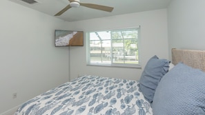Ein Schlafzimmer mit einem Bett, einem Deckenventilator, einem Fenster mit Jalousien und einem an der Wand montierten Fernseher.