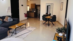 A modern living room with a sofa, coffee table, and a dining area with chairs and a table. There is a kitchen in the background, a fire extinguisher on the wall, and a flat-screen TV mounted on the wall. - St. Julian's Central Apartment 5 (St. Julian's)