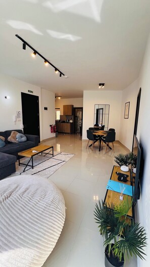 Ein modernes Wohnzimmer mit Sofa, Couchtisch und einem Essbereich mit Stühlen und Tisch. Im Hintergrund befindet sich eine Küche, an der Wand ein Feuerlöscher und ein Flachbildfernseher.