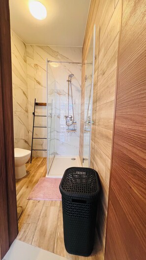 Ein Badezimmer mit Duschkabine aus Glas, Holzboden, schwarzem Mülleimer und weißer Toilette.