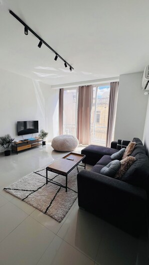 Un saló modern amb televisió plana, un sofà, una taula de centre i una finestra gran amb cortines.