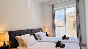 A hotel room with a large bed, a bedside table with a lamp, a window with curtains, and an air conditioning unit. - St. Julian's Central Apartment 5 (St. Julian's)