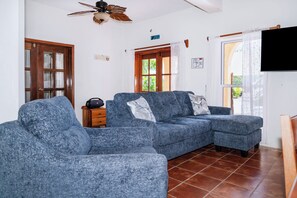 Ein Wohnzimmer mit einem Ecksofa, einem Deckenventilator, einer Holztür und einem Fenster mit Vorhängen.