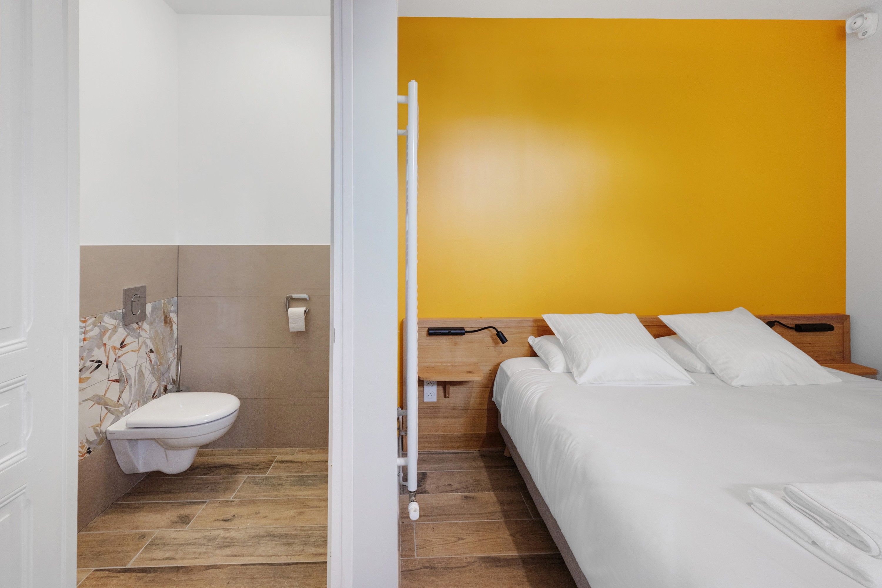 Una habitación de hotel con piso de madera, una cama blanca y una pared de acento amarilla.