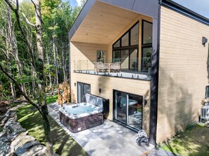 A modern two-story house with a hot tub, outdoor seating, and a large window overlooking a forested area.