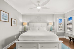 Une chambre à coucher comprenant un lit, des tables de chevet, un ventilateur de plafond et une fenêtre avec des stores.