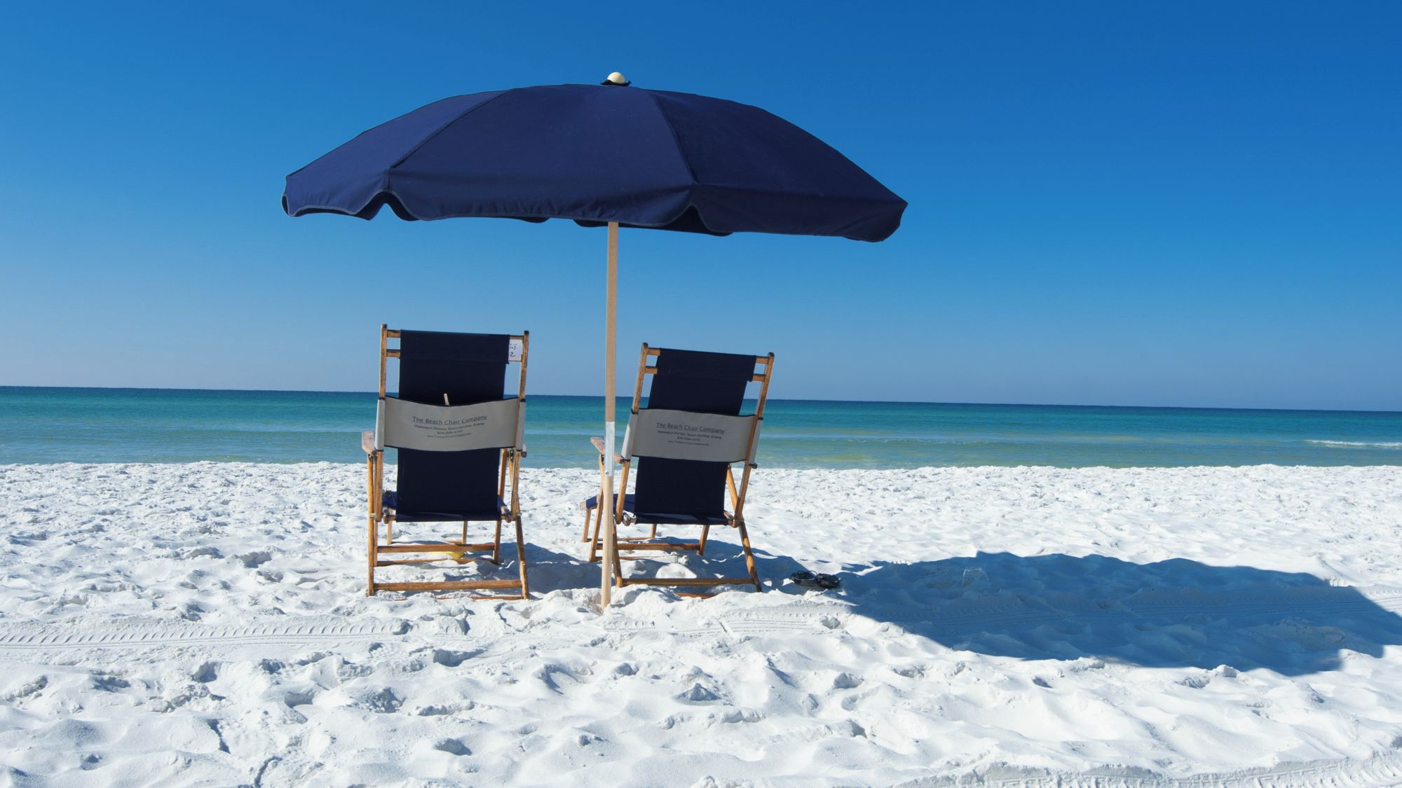Deux chaises de plage, avec un parasol bleu, sont installées sur une plage de sable fin, et on aperçoit de l’eau d’un bleu limpide à l’arrière-plan.