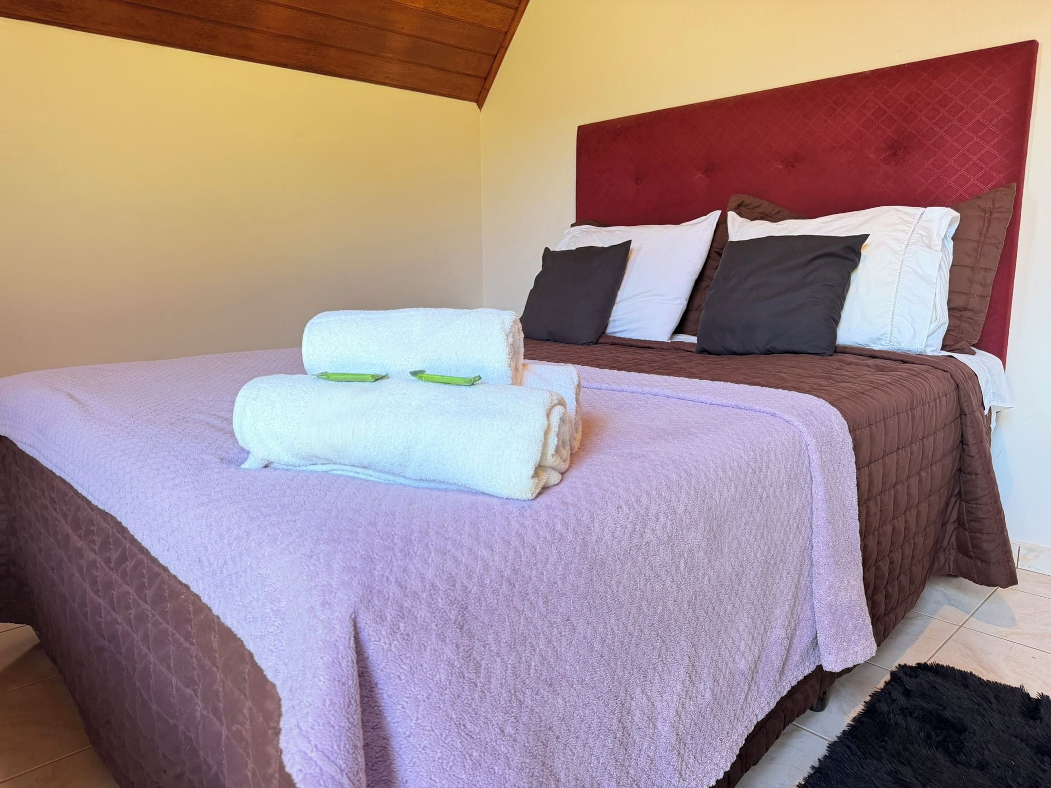 A neatly made bed with a purple blanket and two rolled towels, a red headboard, and a brown quilted bedspread.