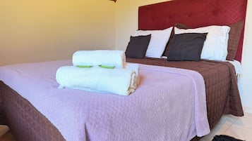 A neatly made bed with a purple blanket and two rolled towels, a red headboard, and a brown quilted bedspread.