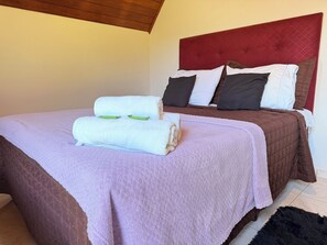 Una cama bien tendida con una colcha morada y dos toallas enrolladas, cabecera roja y edredón acolchado marrón.
