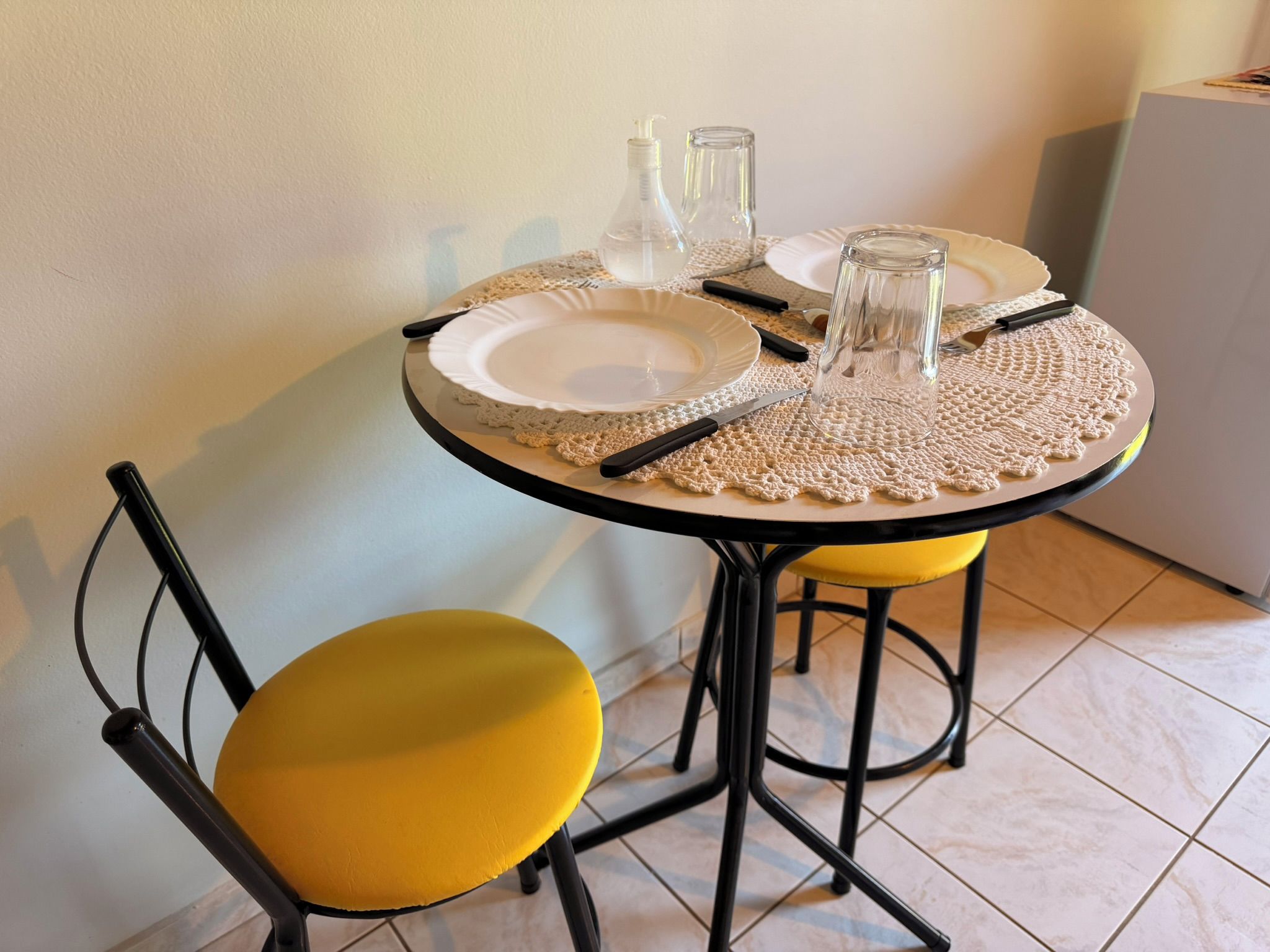 A round dining table with a white plate, two glass bottles, and a glass. There are two yellow chairs with black frames.