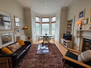 Un salon avec un canapé noir, un tapis à motifs, une table à manger avec des chaises, une cheminée et des tableaux encadrés accrochés aux murs.