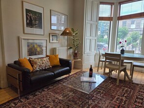 Ein Wohnzimmer mit einem schwarzen Ledersofa, einem Glastisch, einem Holzstuhl und einem gemusterten Teppich. An den Wänden hängen gerahmte Bilder und es steht ein Blumentopf bereit.