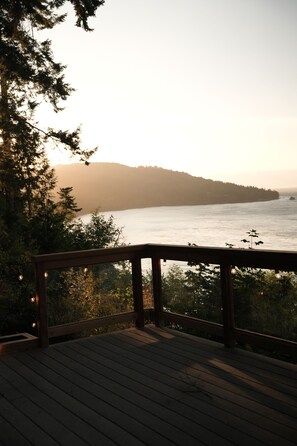 A wooden deck with a railing overlooking a body of water and a forested hill in the distance. - Dreamy Castle in the woods with ocean views, hot tub, private gazebo & glowing sunsets—just minutes from Olympic National Park. (Port Angeles)