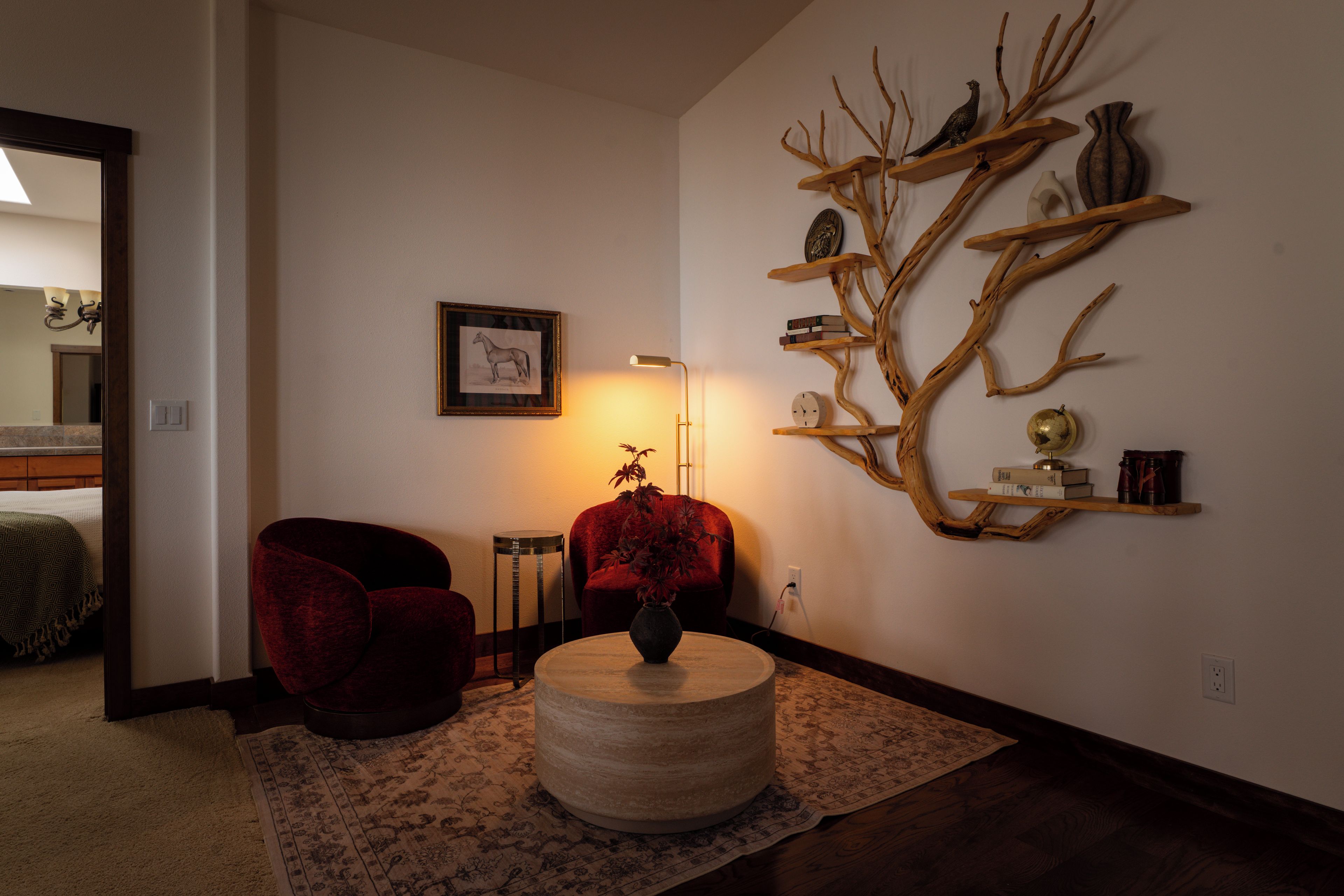 A living room with a red armchair, a round coffee table, a wall-mounted wooden tree sculpture, and a framed picture.