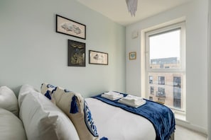 Een slaapkamer met een groot raam, een bed met wit en blauw beddengoed en drie ingelijste kunstwerken aan de muur.