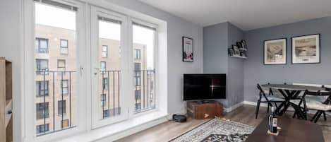 Ein modernes Wohnzimmer mit großem Fenster, Flachbildfernseher, Esstisch mit Stühlen und einem gemusterten Teppich.