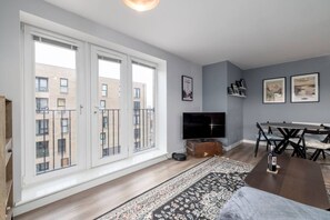 Een moderne woonkamer met een groot raam, een flatscreen televisie, een eettafel met stoelen en een kamerkleed met een patroon.