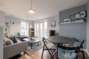 Een moderne woonkamer met een grijze bank, een ronde eettafel en een salontafel. Er zijn twee hanglampen, een boekenkast en een open haard.