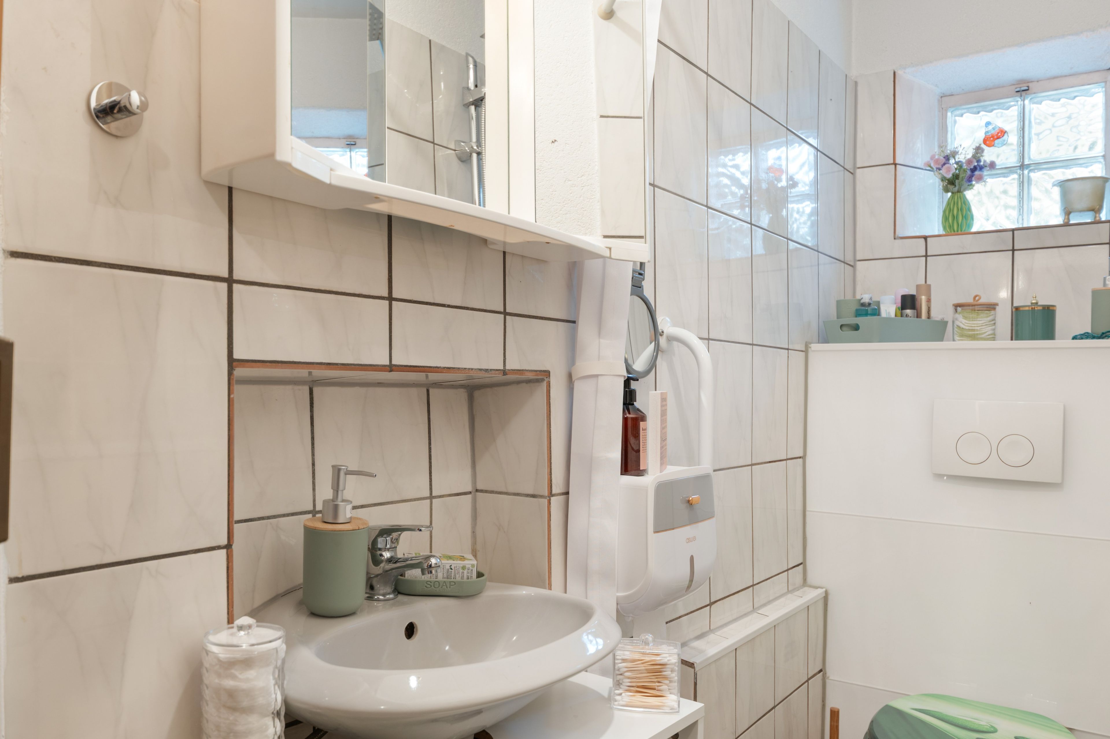 Ein Badezimmer mit einem weißen Waschbecken, einem Spiegel, einer Dusche und einem Fenster mit Blick auf Bäume.