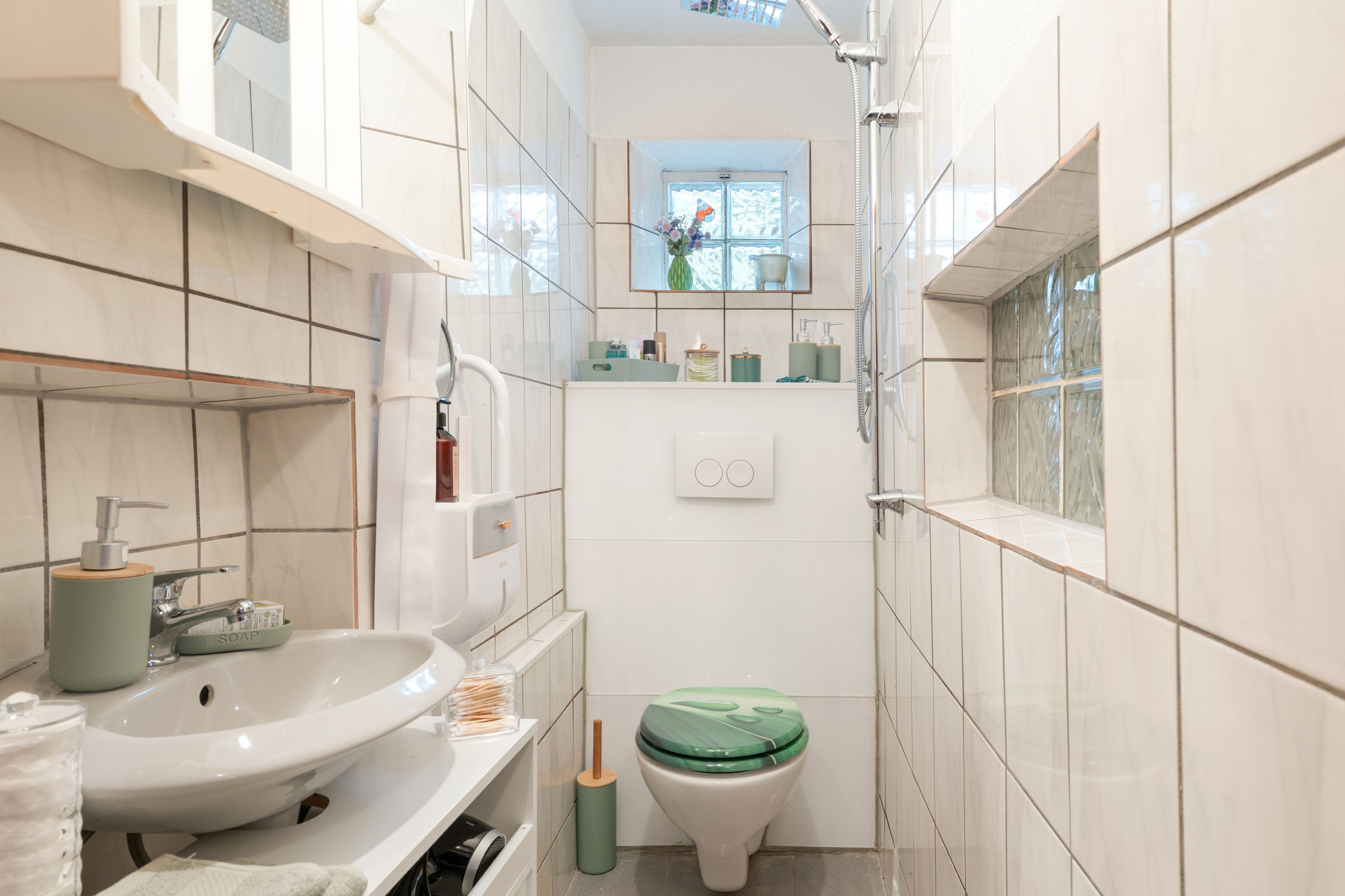 Een kleine badkamer met een witte wastafel, een toilet met een groene deksel, en een raam boven het toilet.