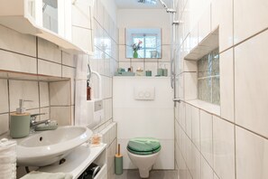 Casa de banho estreita com lavatório branco, sanita com tampa verde e janela sobre a sanita.