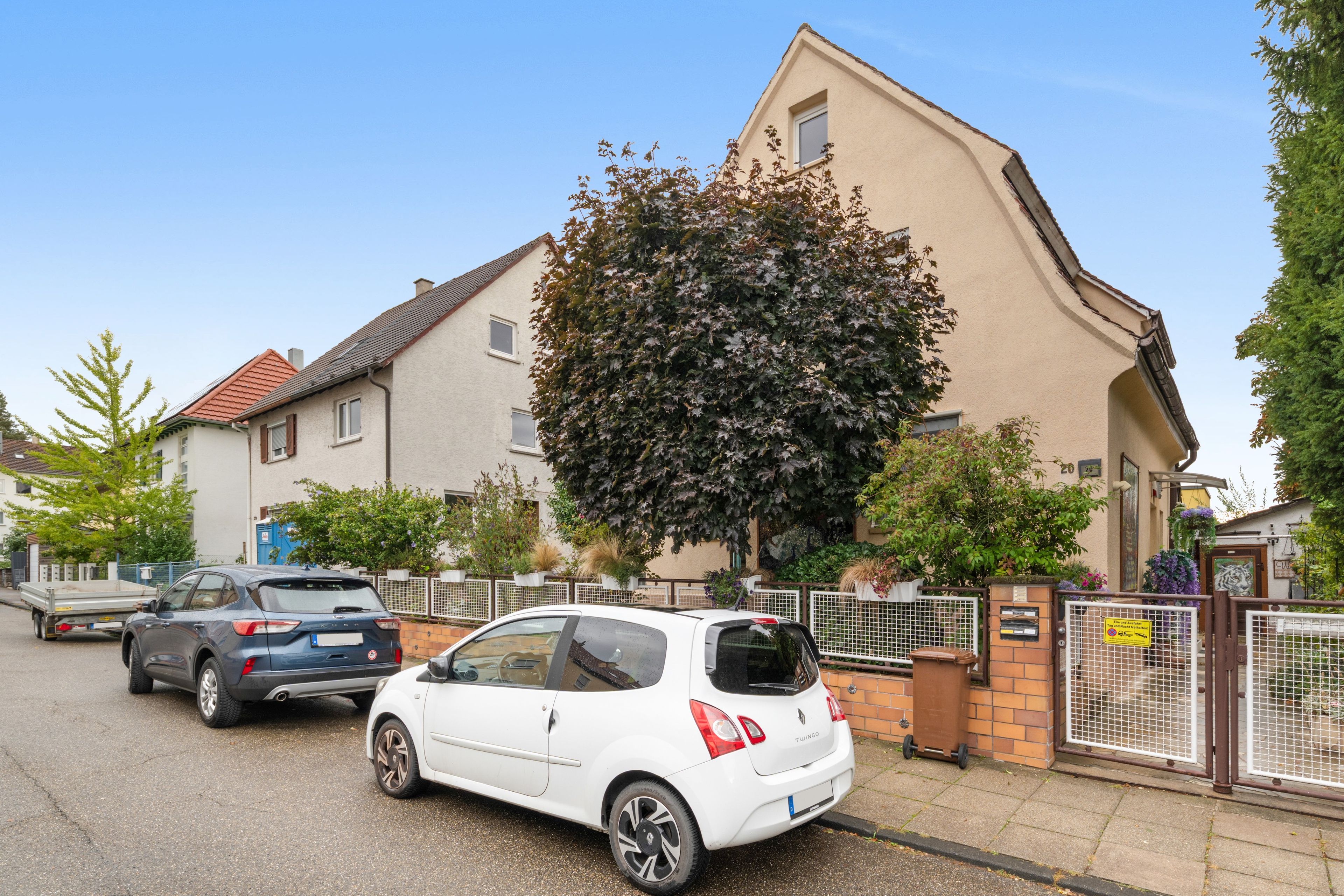 A residential area with two parked cars, a house with a sloped roof, and a large tree in front.