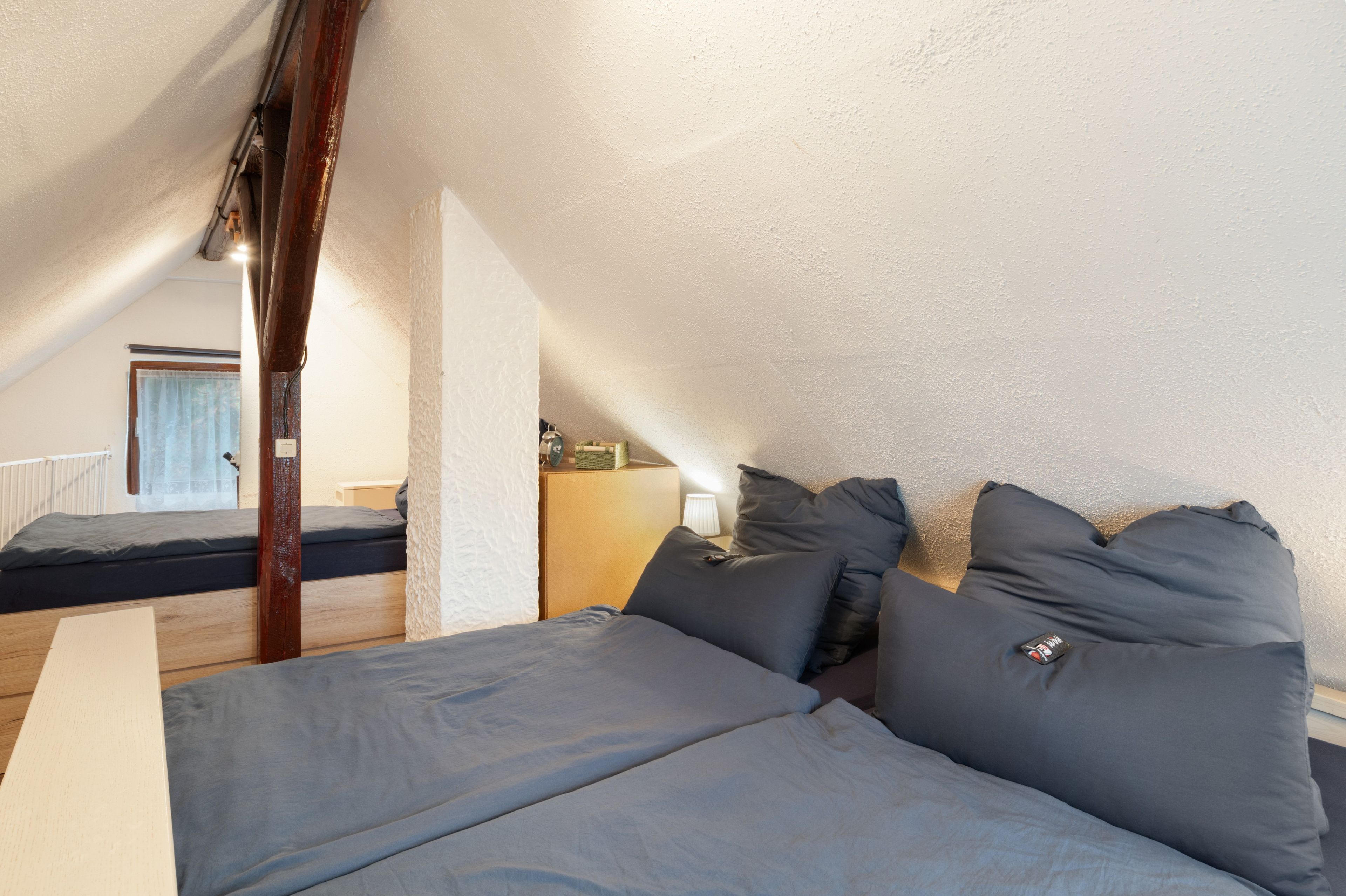 Ein Schlafzimmer mit schrägem Dach, einem Holzbalken, einem Bett mit grauer Bettwäsche, einem Nachttisch und einer Lampe.