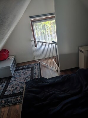 Een kamer met een schuine plafond, een raam met uitzicht op groen, een witte radiator en een kamerbreed tapijt met een patroon.